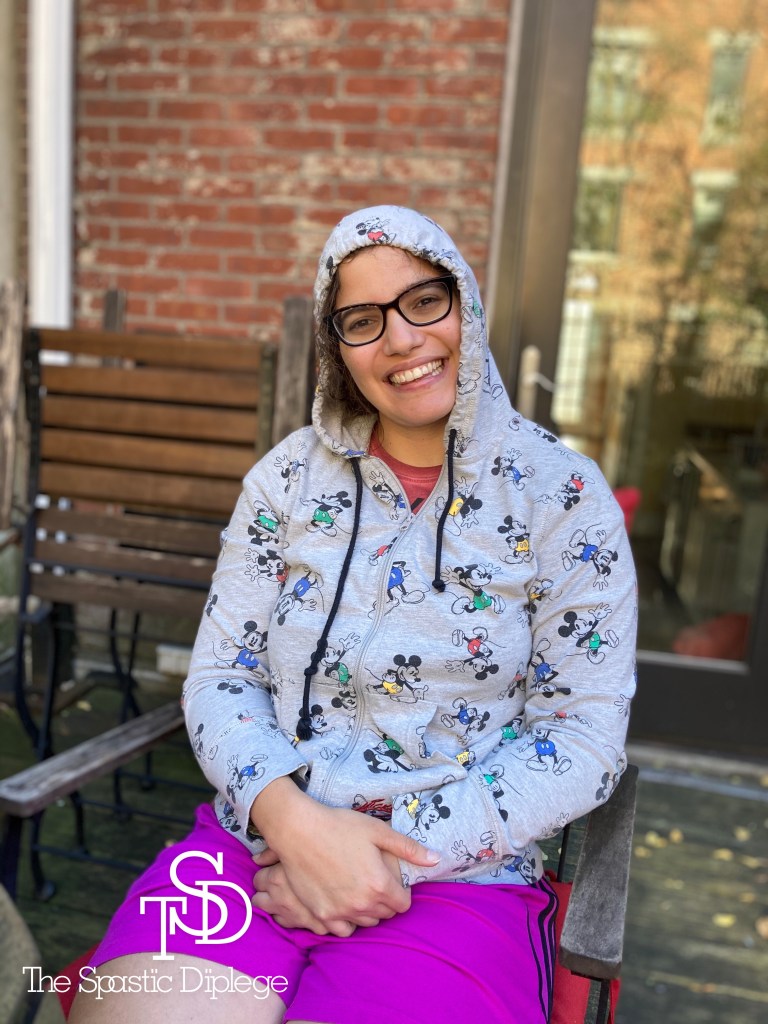 Me sitting on a chair on a deck, wearing a Mickey Mouse zip up hoodie and purple shorts. A door is behind me. I'm smiling with my hands on my lap. My logo is in the bottom hand corner in white.