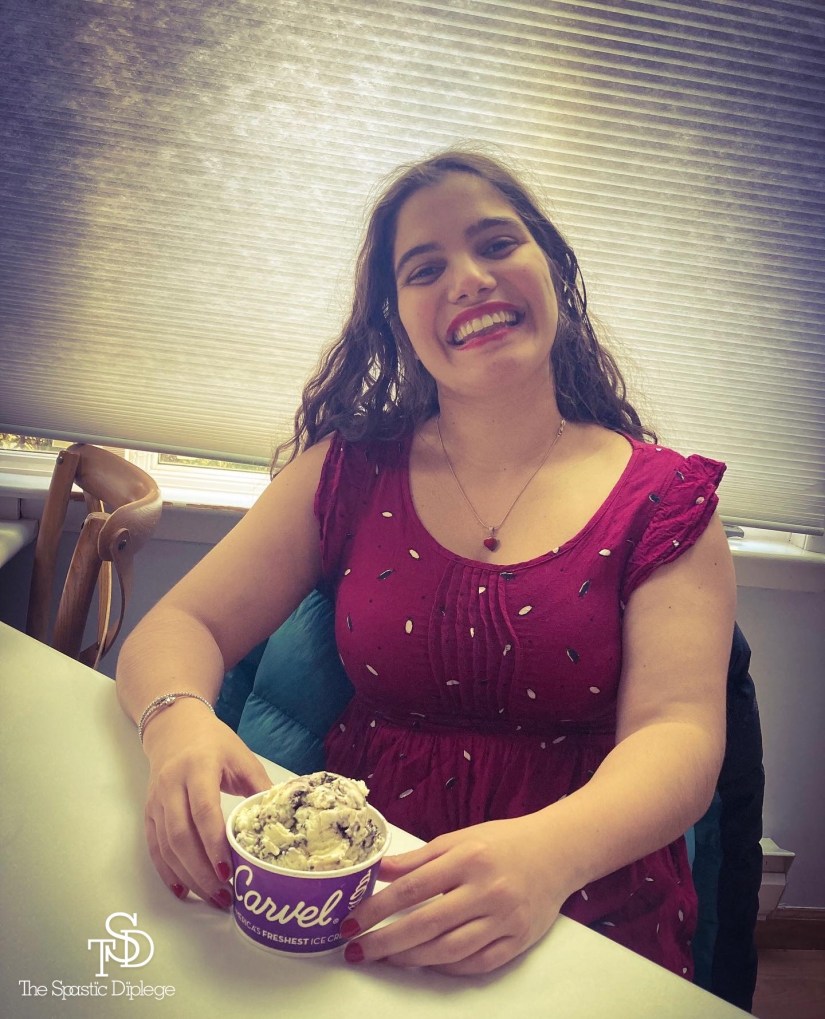 Me sitting down wearing a red dress with a red heart necklace. I'm smiling and posing with a cup of cookies-n-cream ice cream. I'm sitting in front of a window with the blinds down.