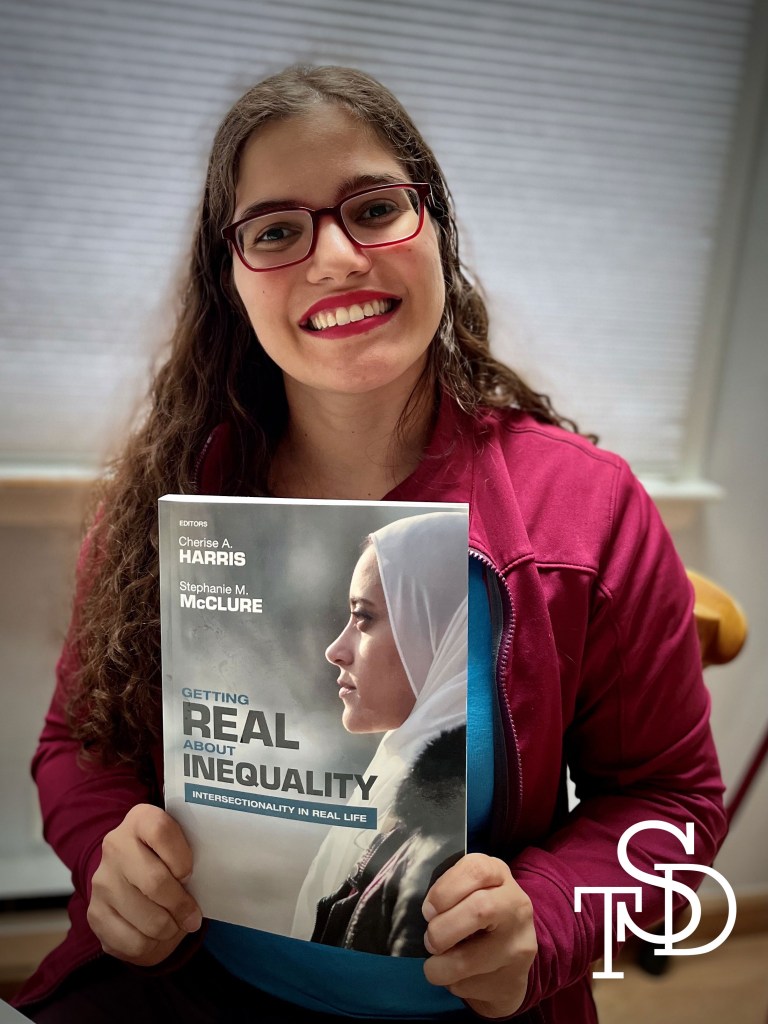 Me sitting and holding the book GETTING REAL ABOUT INEQUALITY: INTERSECTIONALITY IN REAL LIFE. I’m smiling. My logo is in the bottom right corner in white.