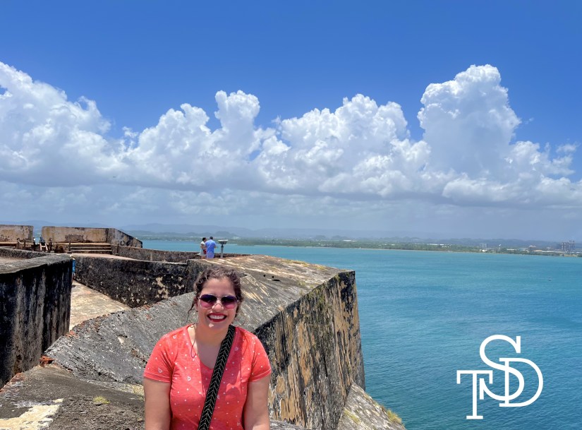 Me standing at El Morro castle in San Juan, Puerto Rico. A big is behind me. I'm smiling and wearing sunglasses and an orange and white polka dot t-shirt. My logo, the letters TSD, is in the bottom right corner in white.