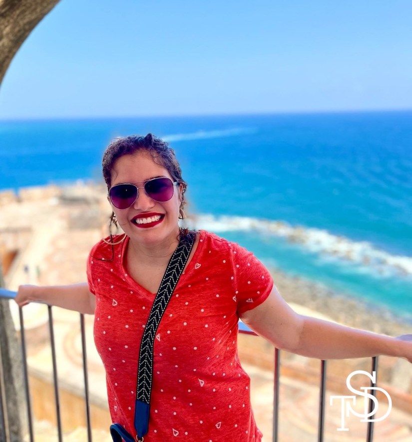 Me, smiling and standing on a balcony, leaning on a railing with the ocean behind me. I am wearing sunglasses and hoop earrings. My logo is in the lower right corner in white.