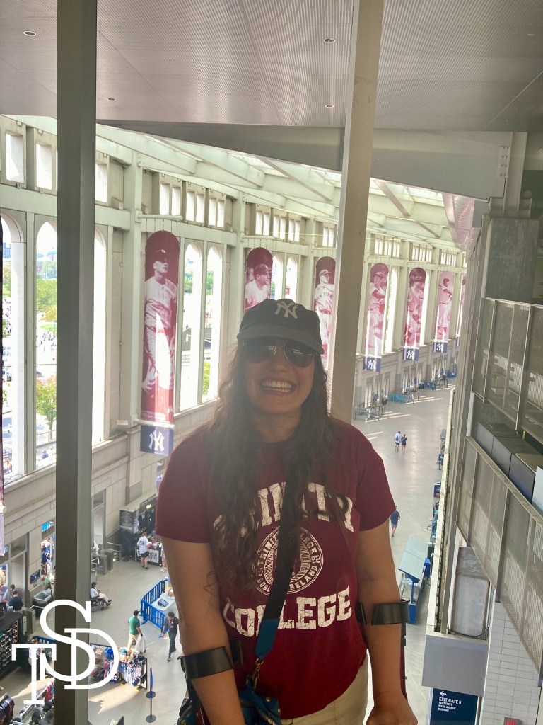 Me standing in Yankee Stadium with banners of famous Yankee legends behind me. I'm wearing a red shirt, a Yankee baseball cap, sunglasses. I'm smiling.
