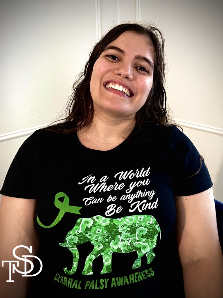 Me, sitting down. I'm smiling and wearing a black tee shirt with white cursive font that reads, "In a world where you can be anything, be kind." to the left of that is a green ribbon for cerebral palsy awareness. There's also a green elephant with the words "cerebral palsy awareness" written underneath it. My logo is in the bottom lefthand corner in white, letters TSD.
