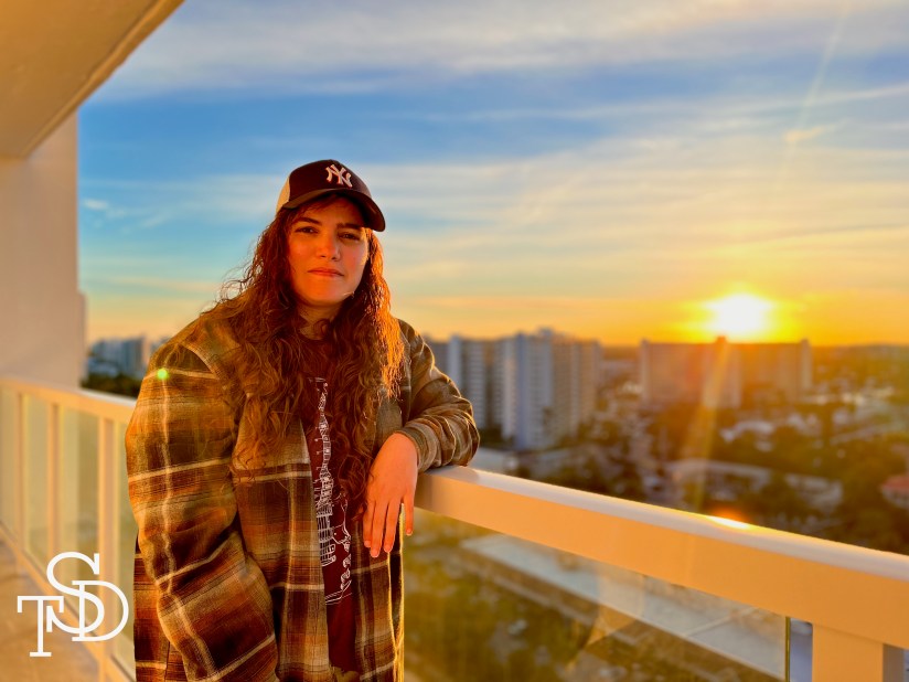 Me, a woman with long, curly brown hair in her late 20s, standing on a balcony with the sun setting behind meI'm wearing an oversized flannel with a red tee shirt underneath, with a Yankee baseball cap.