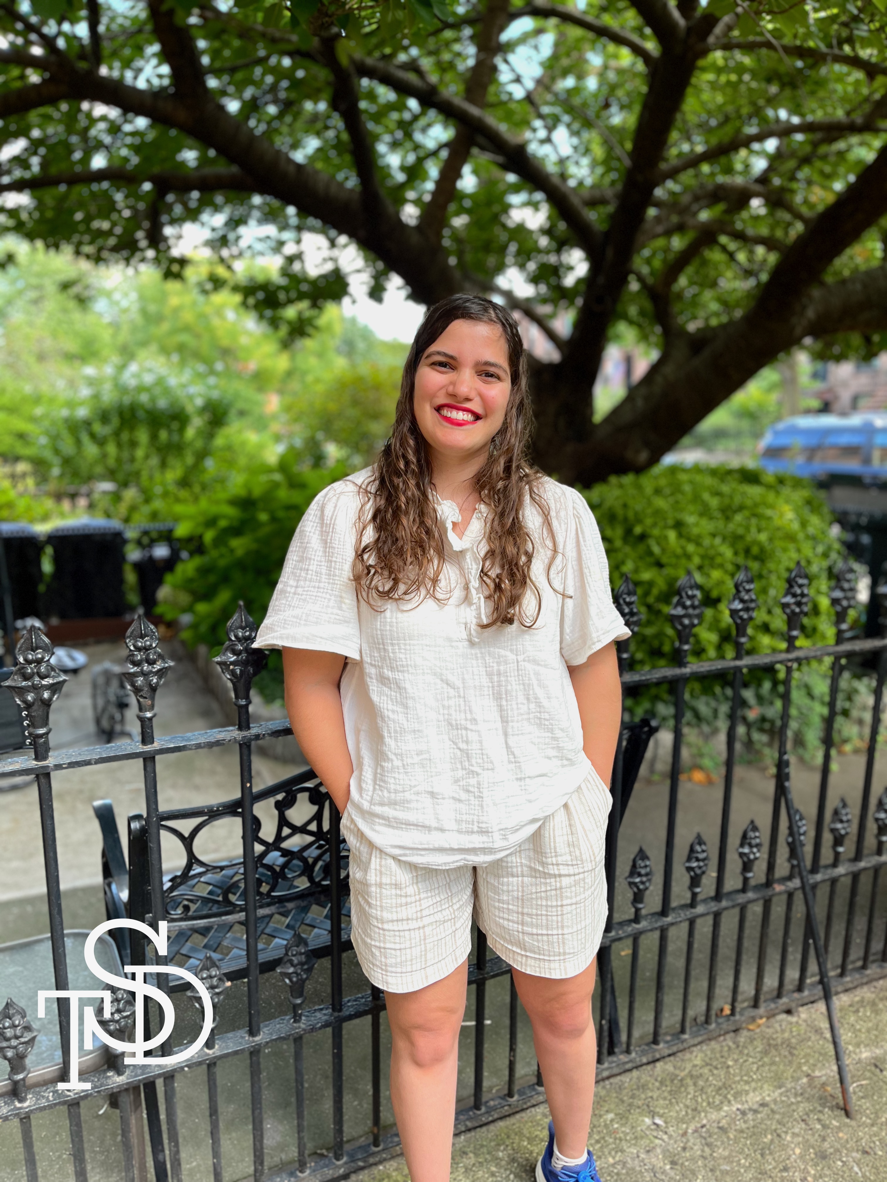 Me, standing against a black fence, with a large tree and greenery in the background. I'm wearing a white blouse and white shorts. I'm smiling.