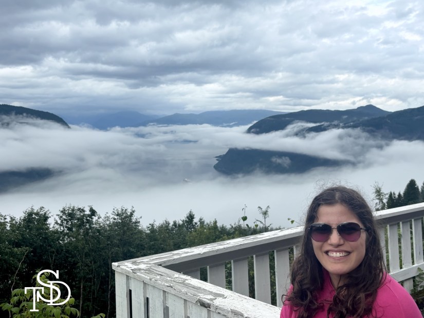 Me standing in front of a backdrops of mountains, water and fog in Norway.  I'm smiling. My logo, the letters T, S, D are in the bottom left hand corner in white.